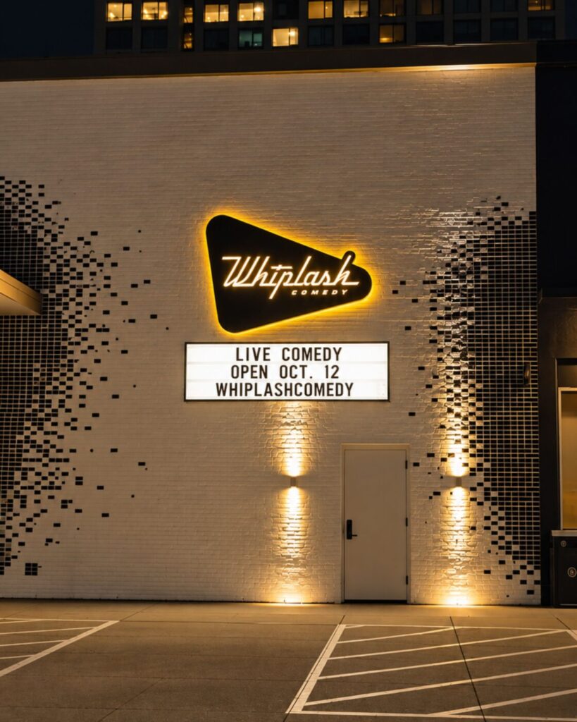 Lit Whiplash Comedy sign on a brick exterior wall with a marquee reading “Live comedy open Oct. 12.”