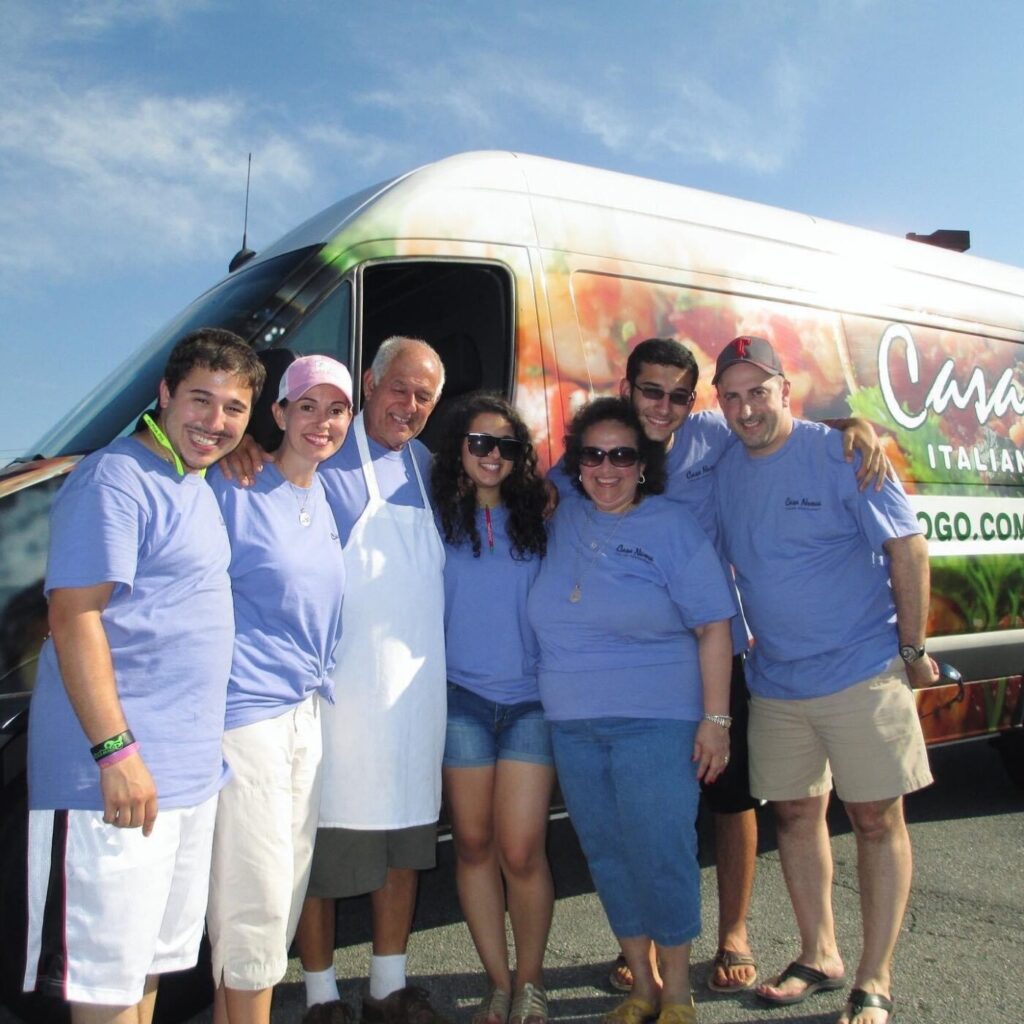 Several people in matching shirts stand beside a branded food truck under a blue sky.