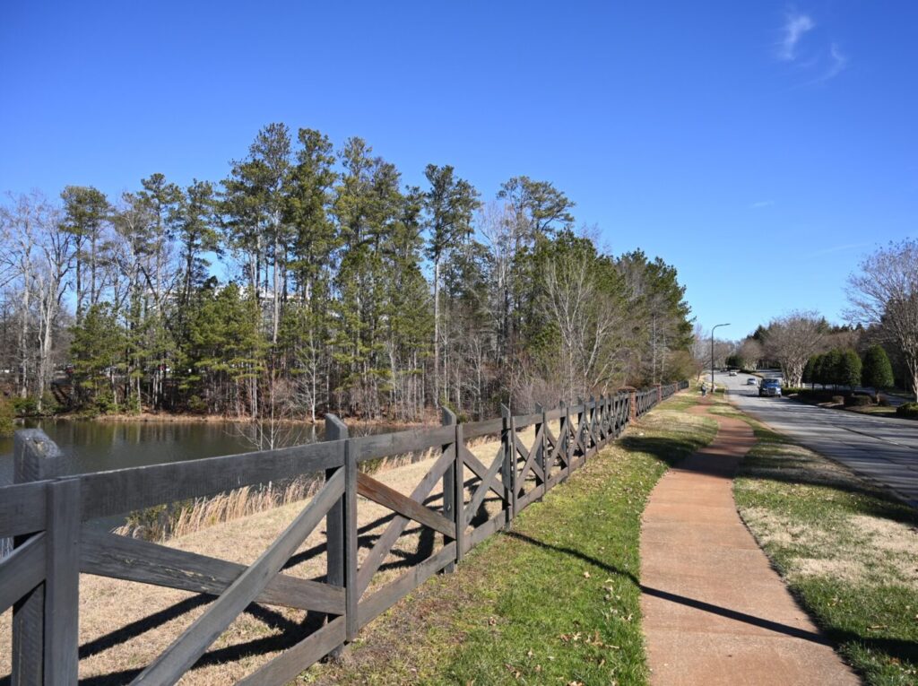 Sidewalk running beside a wooden fence along a road next to a pond and trees