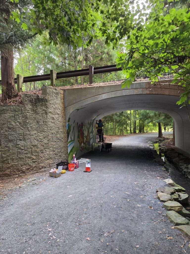 A wide view shows the mural taking shape inside the tunnel with the wooded trail beyond.