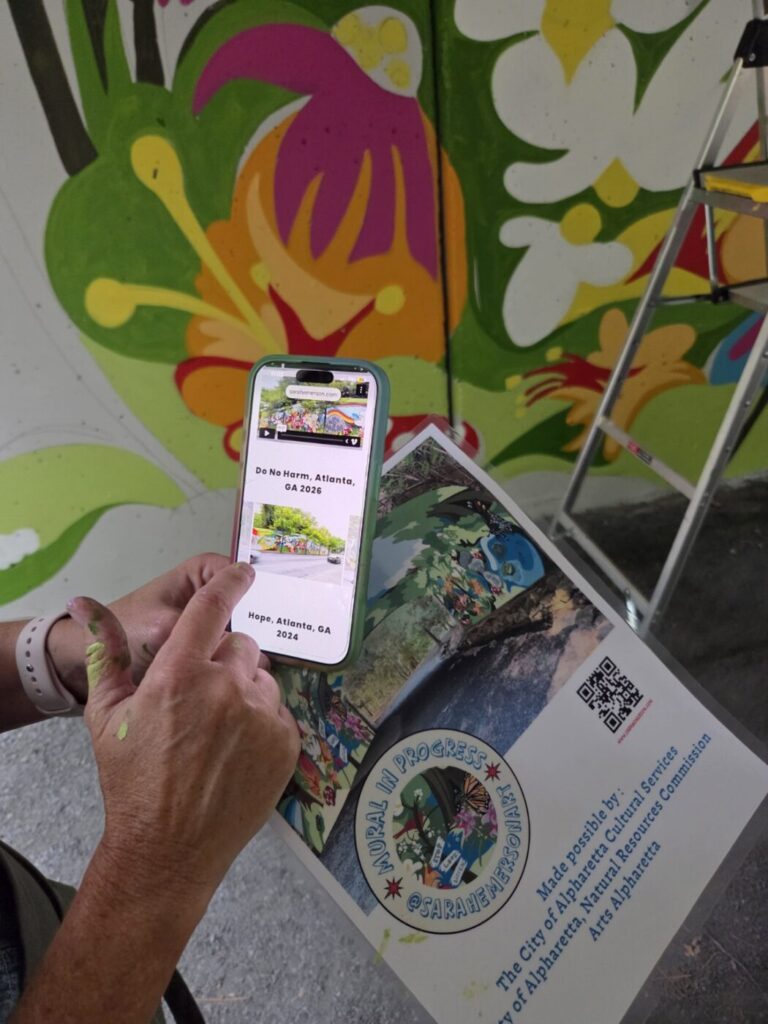 A hand holds a phone showing mural references and a printed design board with project images and a QR code.