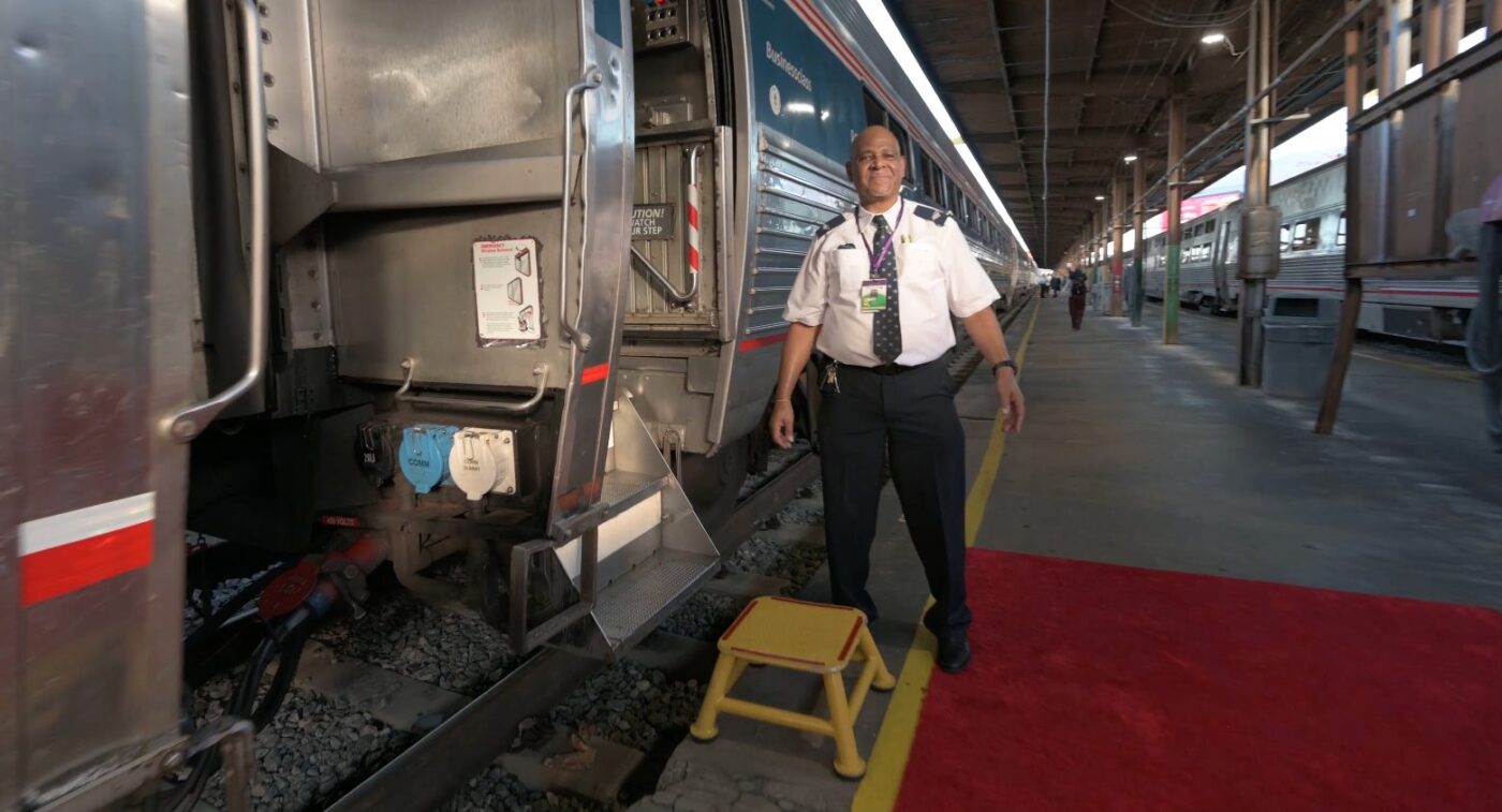 A uniformed Amtrak employee stands on a platform beside an open passenger train door.