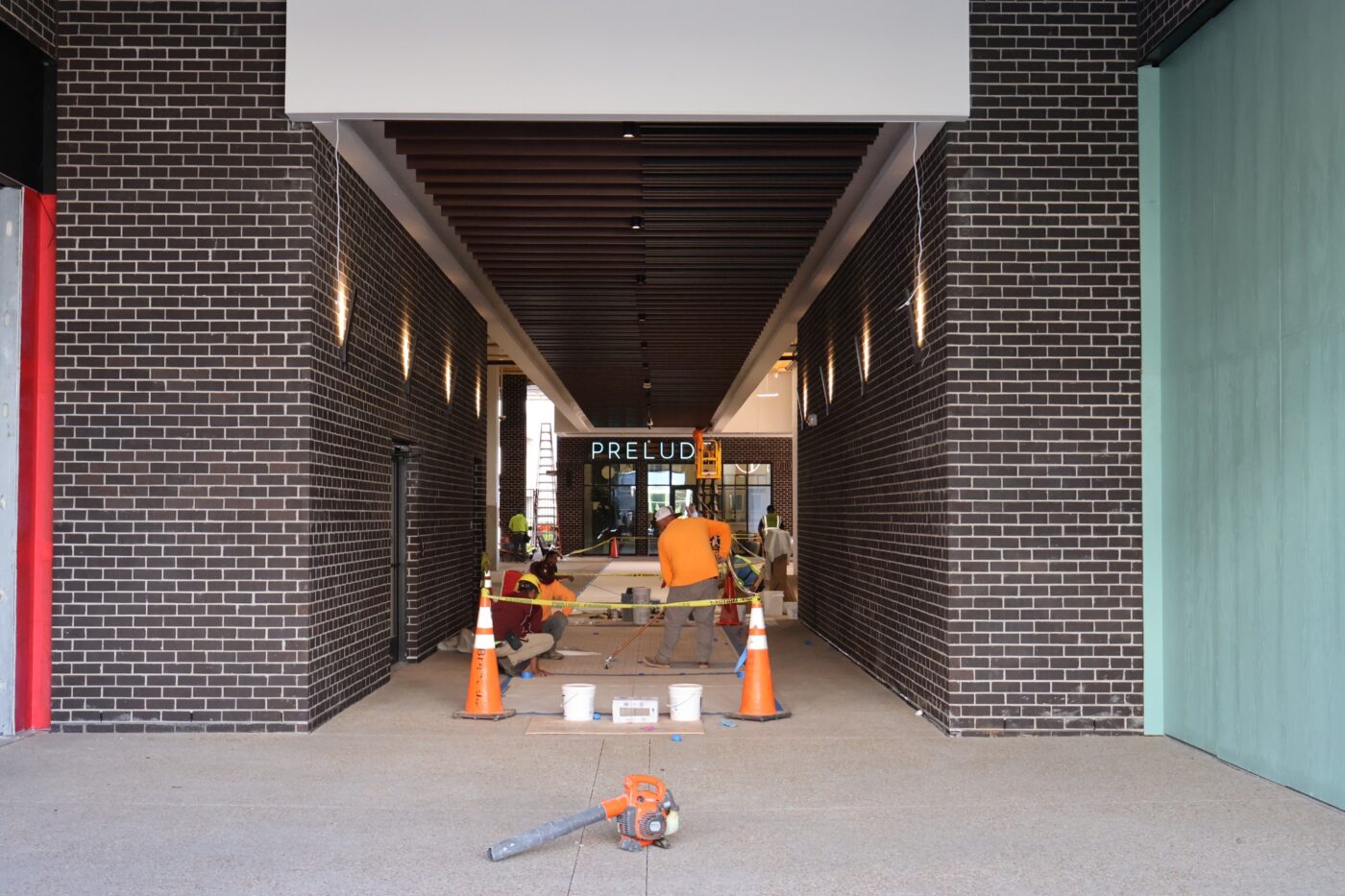 Brick passageway with a Prelude sign at the far end and workers behind a barrier.