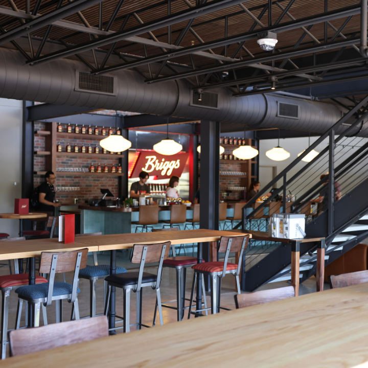 An industrial-style distillery interior with a long wooden bar, hanging lights, exposed ductwork and a staircase beside rows of tables and stools.