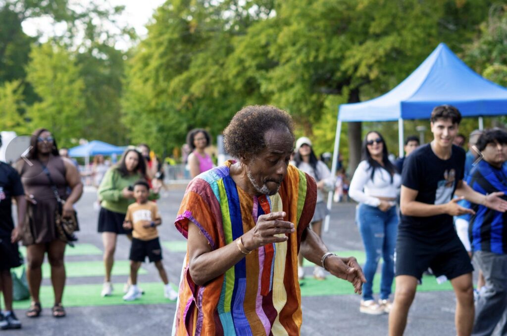 An older man in a colorful striped shirt dances outdoors while festivalgoers watch nearby.