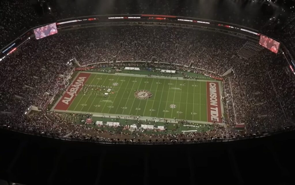 A packed football stadium at night with the field lit and Alabama and Crimson Tide painted in the end zones.