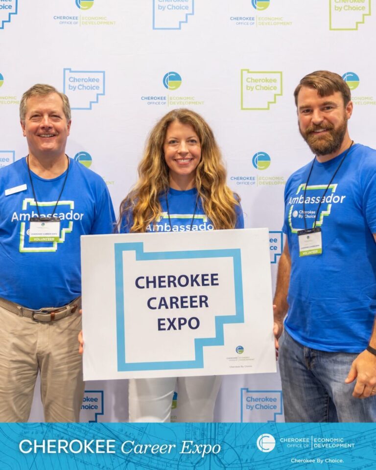 Three volunteers in blue shirts stand behind a sign that reads Cherokee Career Expo.