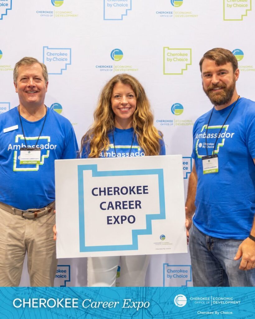 Three volunteers in blue shirts stand behind a sign that reads Cherokee Career Expo.
