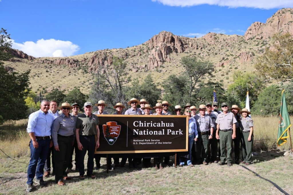 Officials and park employees gather behind the new Chiricahua National Park sign with rocky mountains in the background.