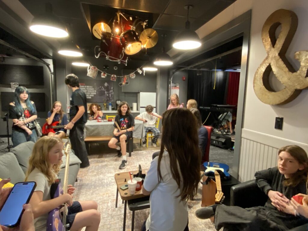 Students gather and talk in a dim rehearsal lounge with drums suspended overhead.