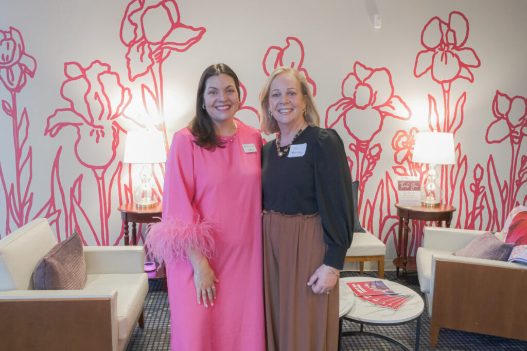 Two women stand smiling in a waiting room with pink floral wall art and lamps behind them.