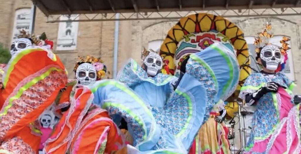 Dancers in skull face paint and bright ruffled dresses perform on an outdoor stage.