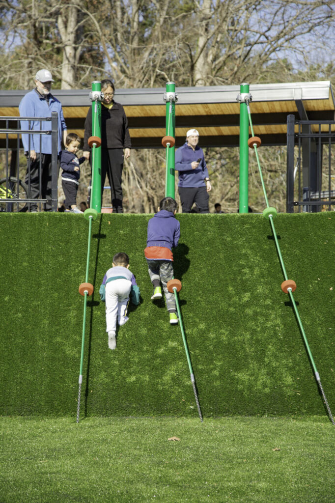 Children climb a steep artificial-grass hill with ropes while adults watch from the top of the play area.