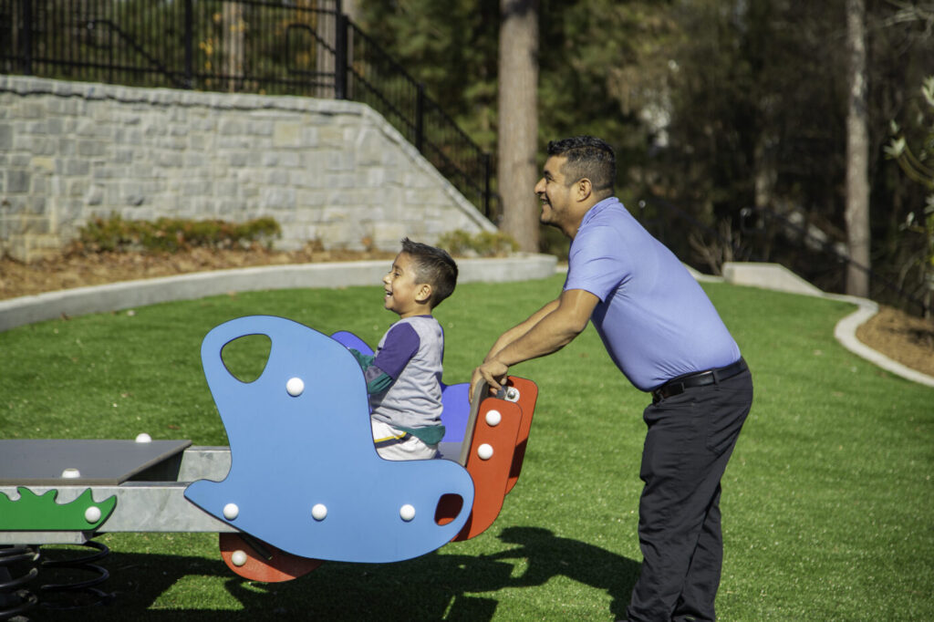 A man pushes a child on a spring rocker in a sunny playground.