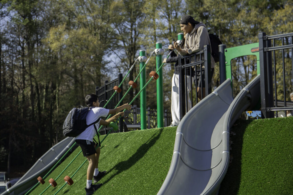 A child climbs a playground ramp beside a slide while an adult stands nearby at the top platform.