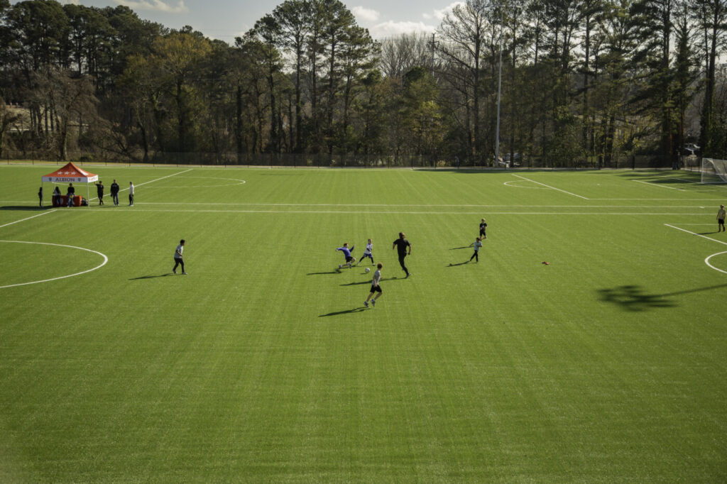 Children and adults play on a wide artificial-turf soccer field with trees beyond the sideline.
