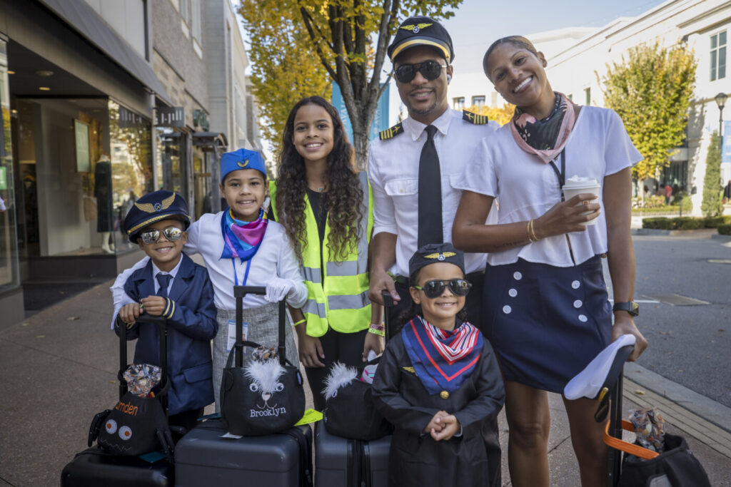 A family of five poses on a shopping district sidewalk, with two children in costumes and a man in a cap and tie
