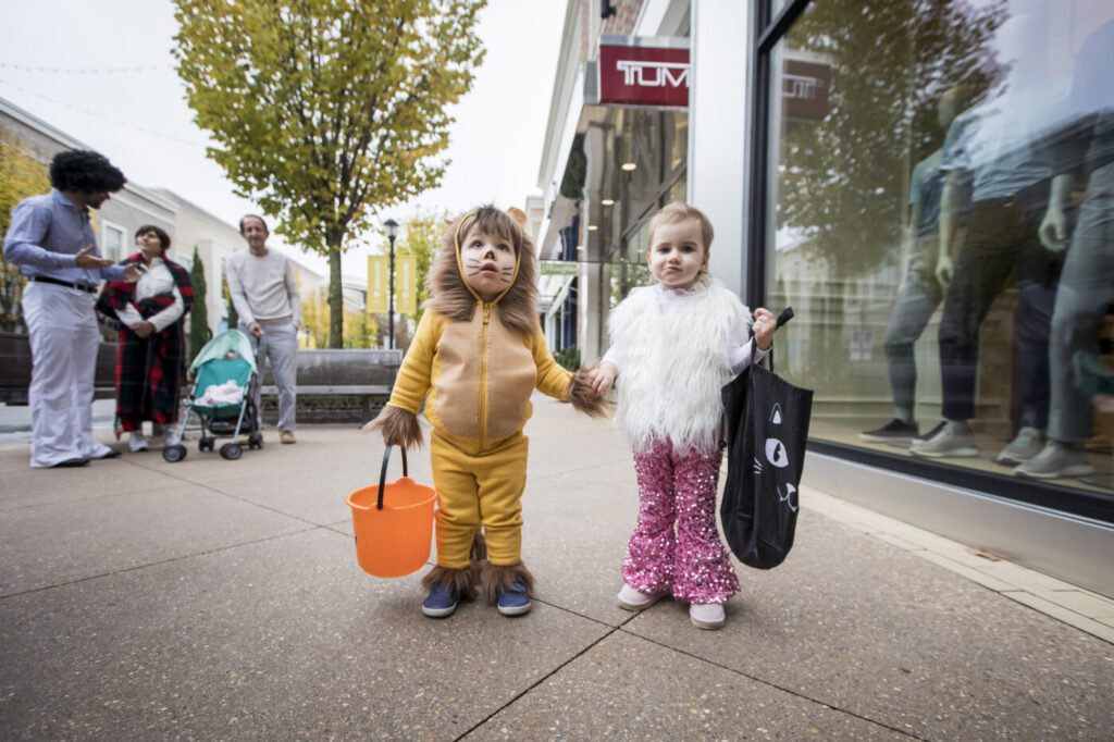 Two children in animal costumes stand hand in hand outside a store, each holding a candy bucket