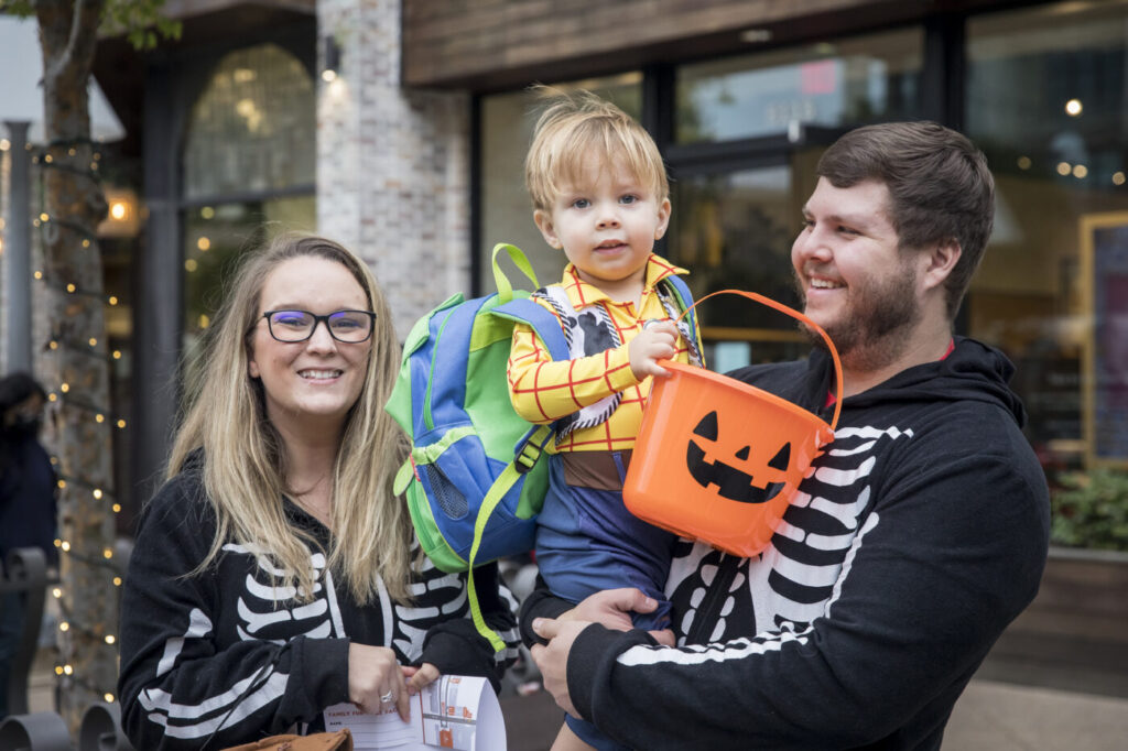 A small child in a skeleton hoodie holds a pumpkin bucket while being held by an adult next to another adult