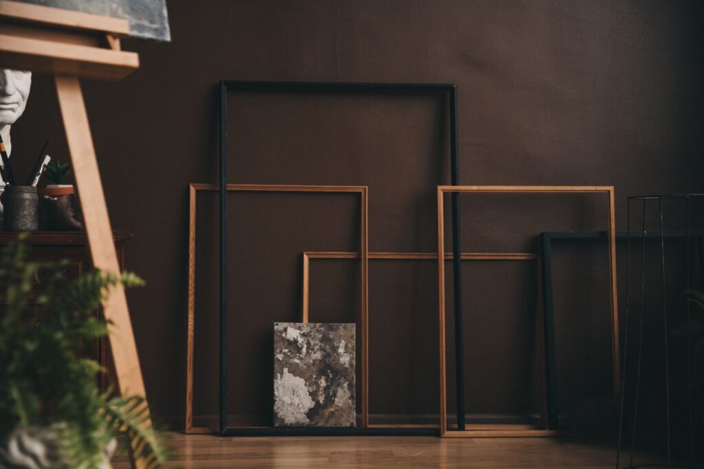 Several empty picture frames of different sizes leaning against a dark wall beside a small art sample and a blurred plant in the foreground.
