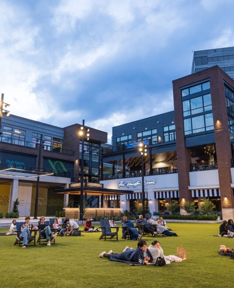 People sit and lie on a grassy plaza surrounded by brick and glass buildings at High Street at dusk.