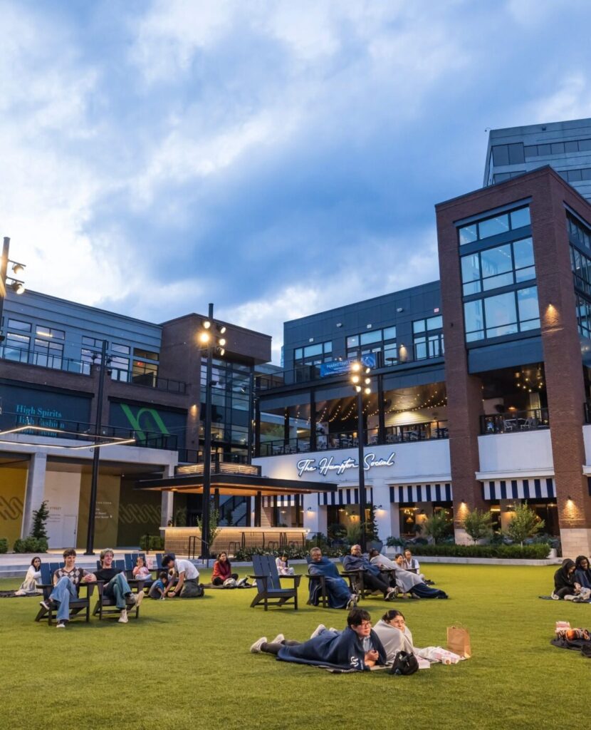 People sit and lie on a grassy plaza surrounded by brick and glass buildings at High Street at dusk.