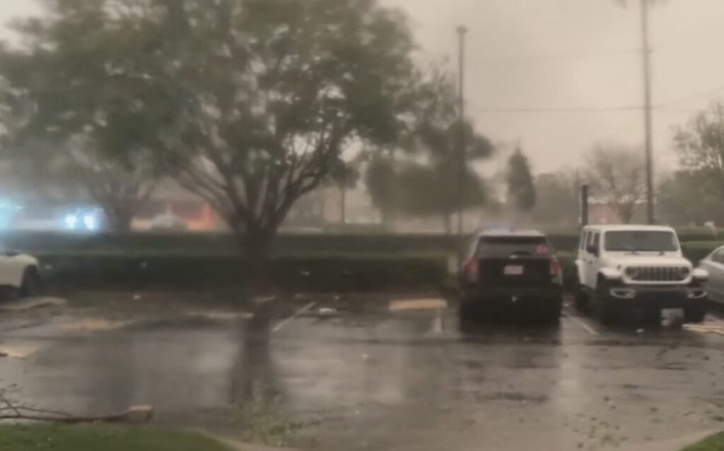 Rain falls over a flooded parking lot with parked vehicles and trees in the background.