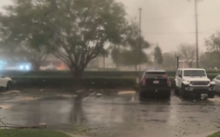 Rain falls over a flooded parking lot with parked vehicles and trees in the background.