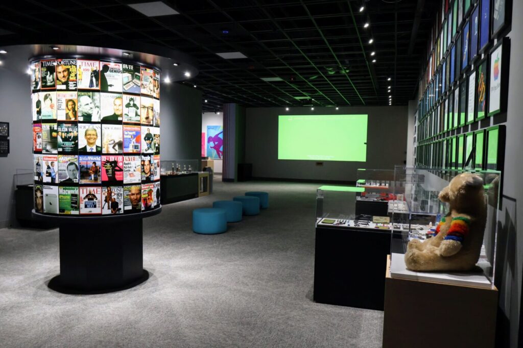 A cylindrical display of magazine covers stands in a gallery with a bright green screen at the far end.