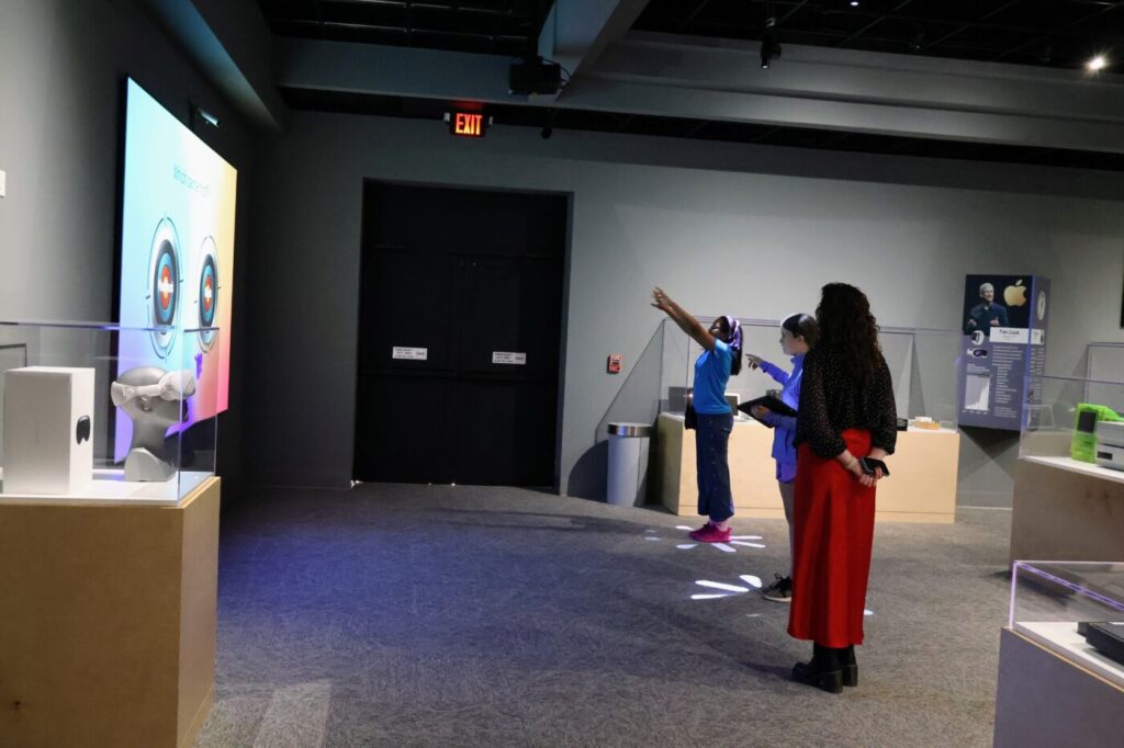 Three visitors stand near a wall with bright graphics projected on it.