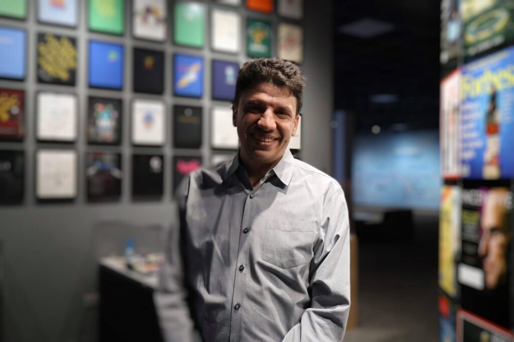 A smiling man stands in a museum gallery with framed magazine covers behind him.