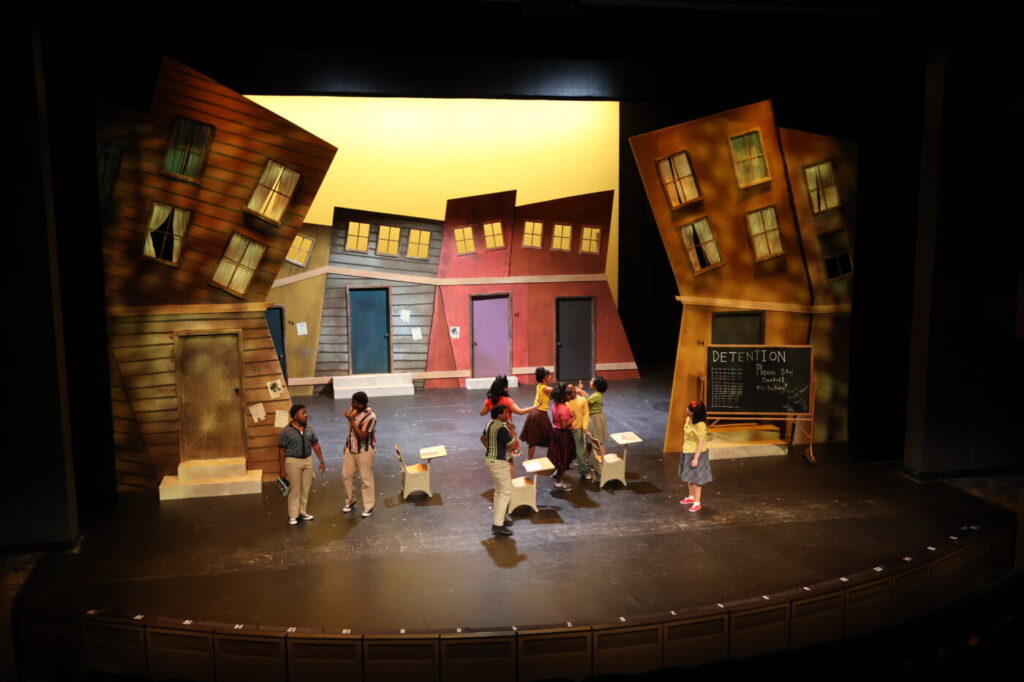 A cast of performers dances on a brightly lit stage surrounded by tilted house facades and a large soda bottle set piece.