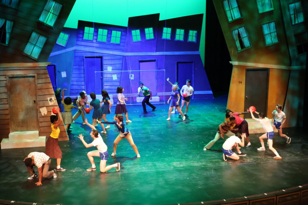 Performers gather on a stage set with tilted houses and a chalkboard labeled Detention.