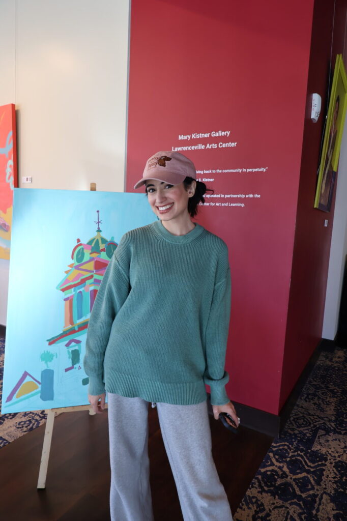 A woman in a pink cap and oversized green sweater stands smiling indoors beside colorful artwork.