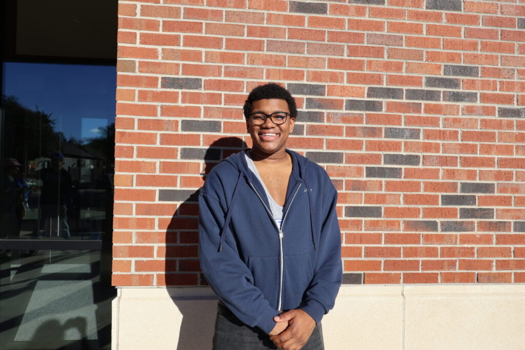 A young man in glasses smiles in front of a brick wall in sunlight.