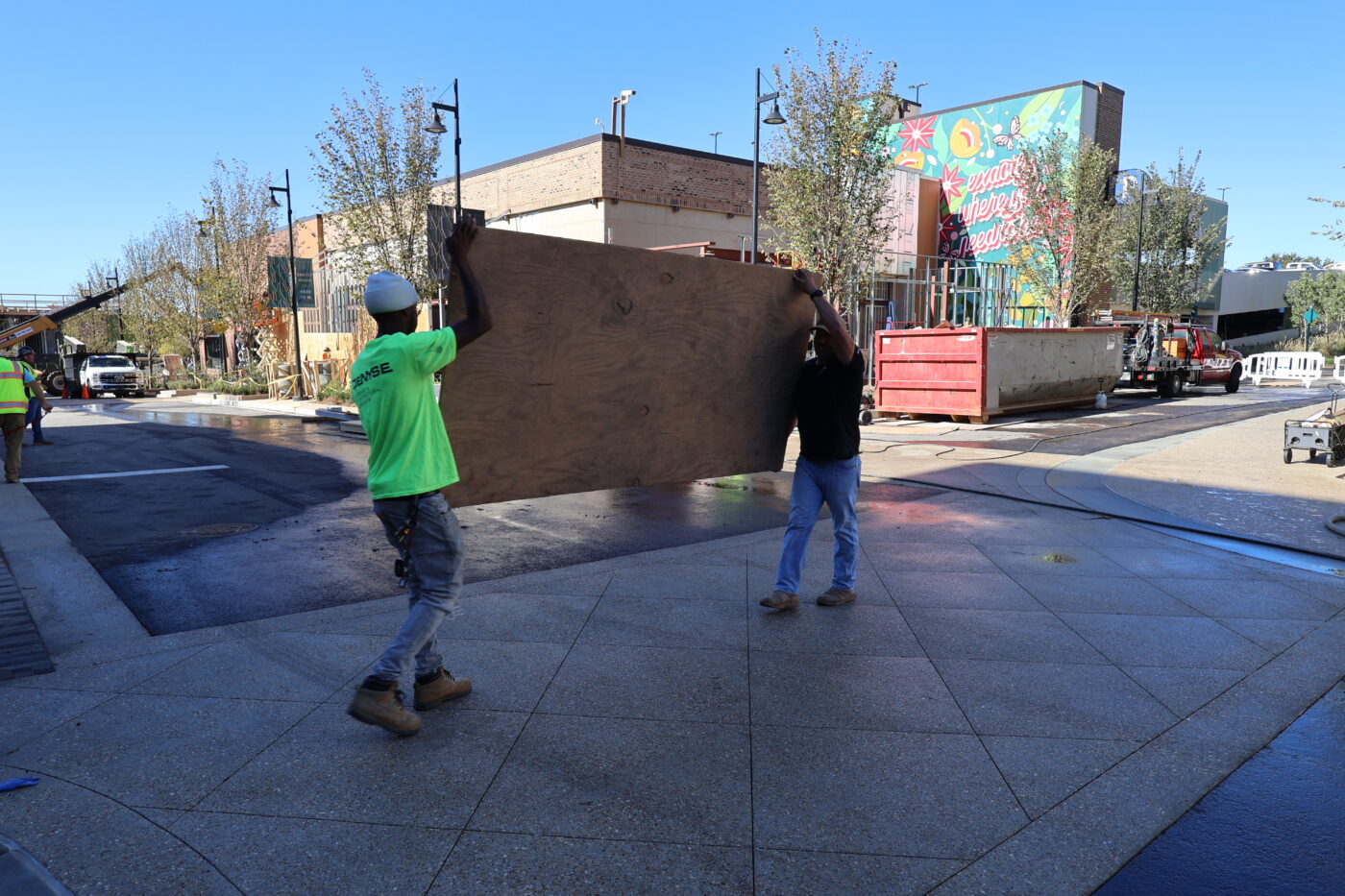 Workers carry plywood through Medley Two workers carry a sheet of plywood across a Medley street, with a colorful mural and construction equipment behind them.