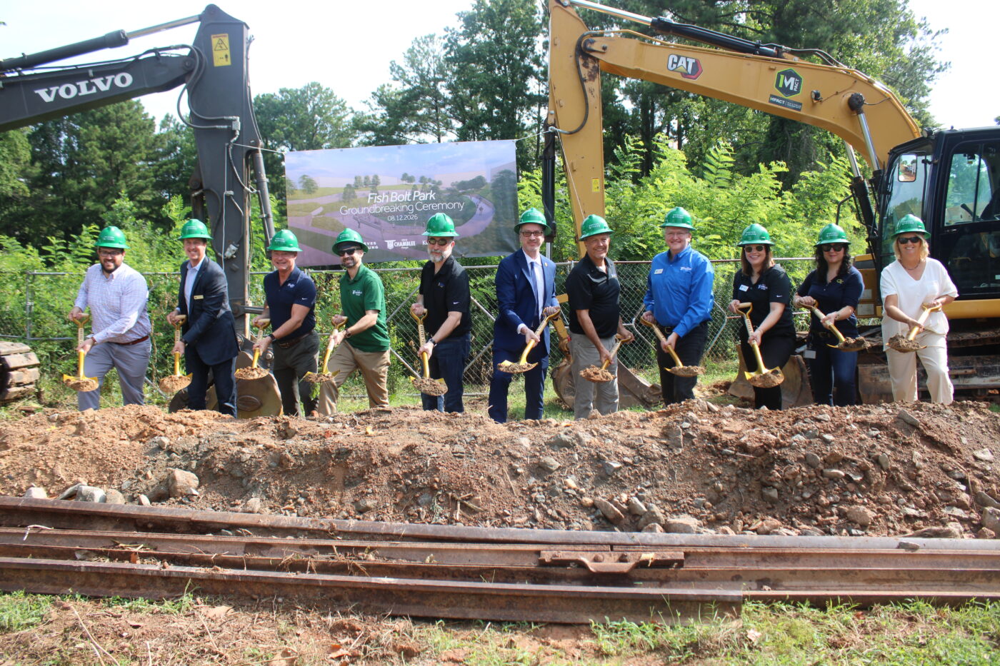 A line of people in green hard hats pose with shovels between two excavators at a groundbreaking site.