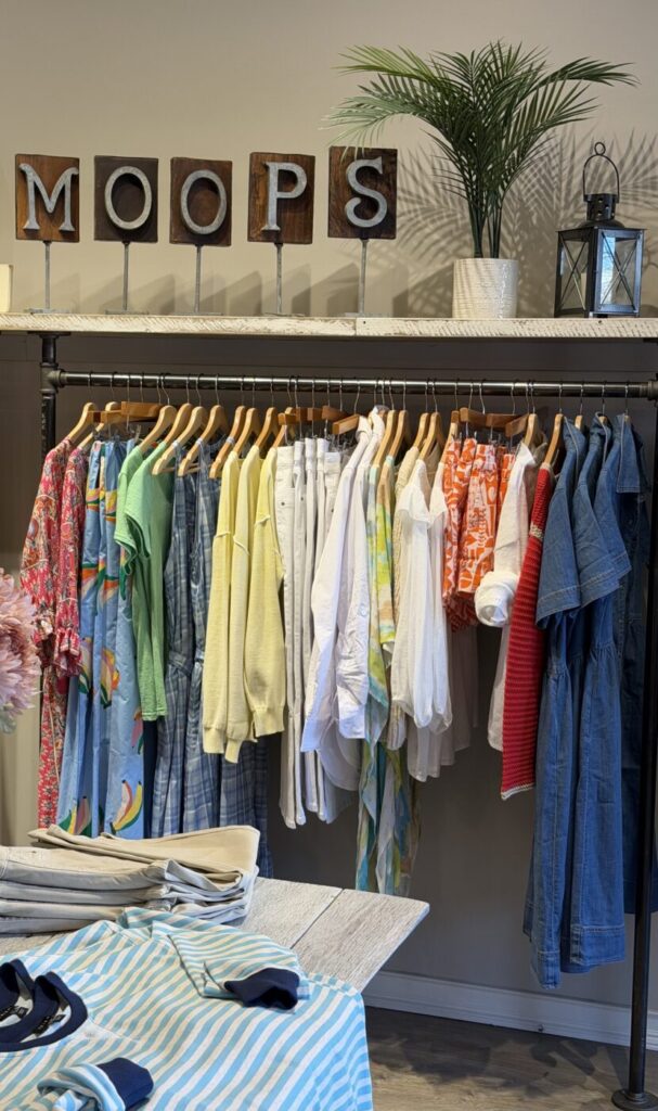 A clothing rack with colorful tops, dresses and jeans beneath a Moop's sign and decorative plant.