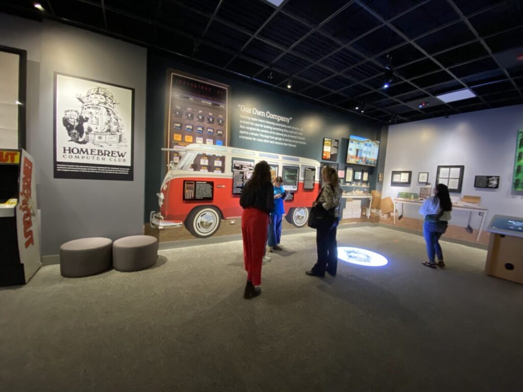Visitors stand in a gallery with a large Homebrew Computer Club poster, a red van display and wall text.
