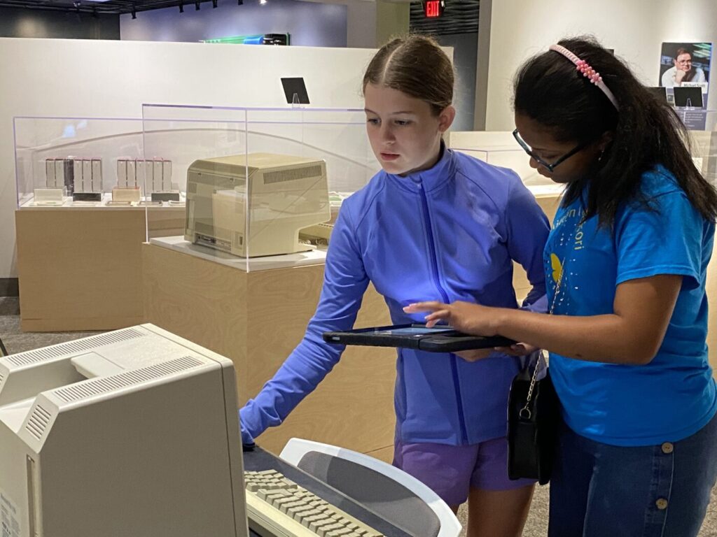 Two girls stand beside a tablet at a museum display with early computer equipment behind glass.