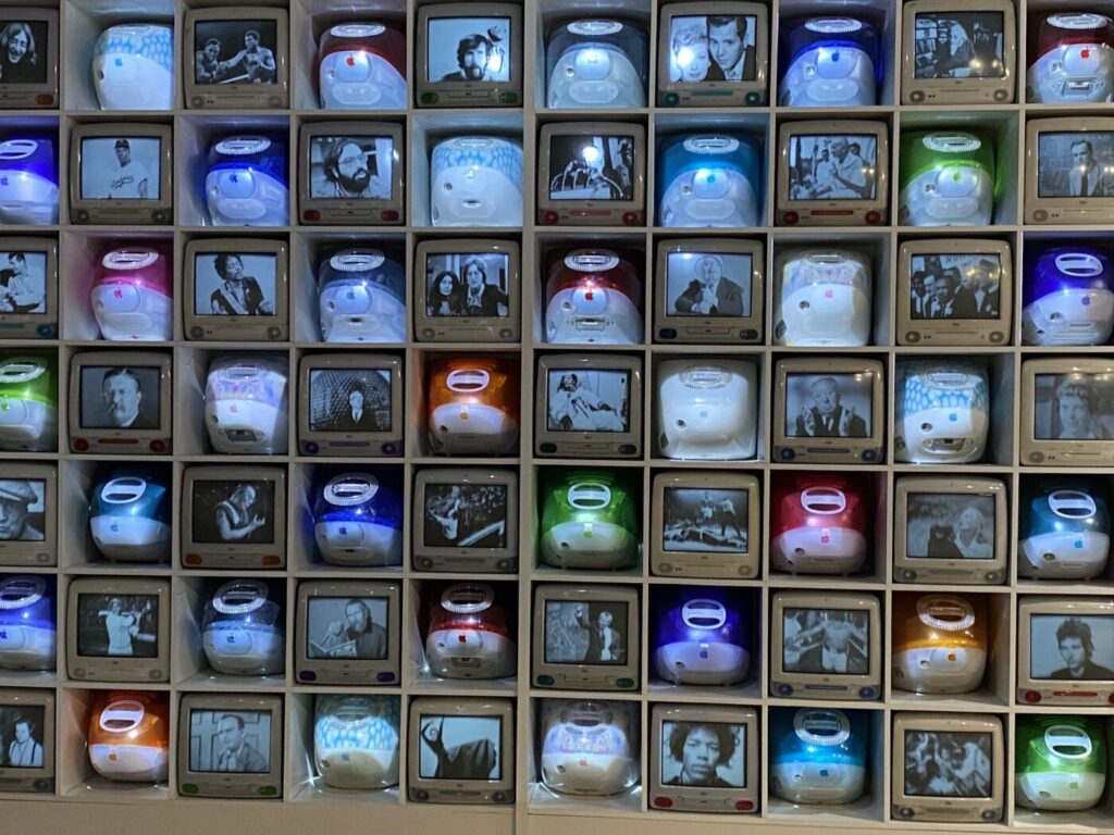 A wall of small screens shows vintage computer images and black-and-white portraits in a grid.