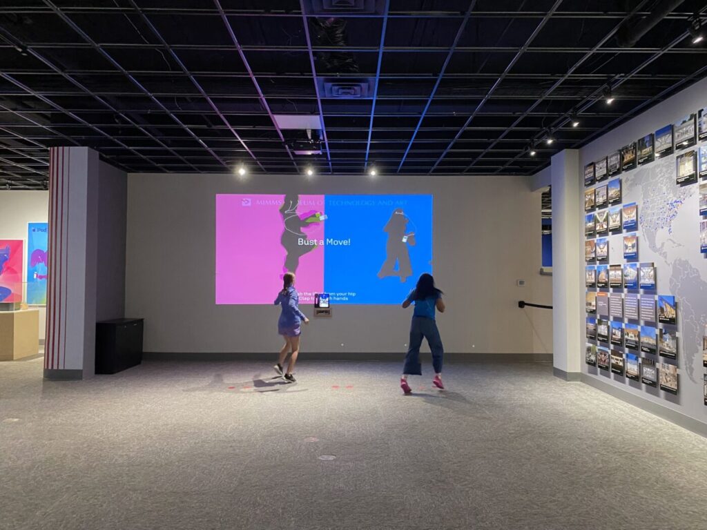 Two visitors dance as their silhouettes appear against pink and blue backgrounds at Mimms Museum’s interactive iPod exhibit.