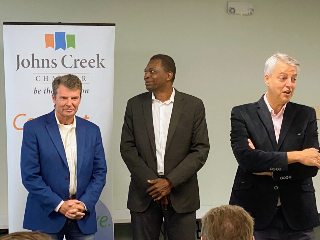Three men stand beside a Johns Creek Chamber banner during an indoor event.