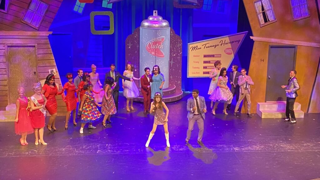 Performers dance on a stage with a large soda-bottle set piece and bright neon-colored backdrops.