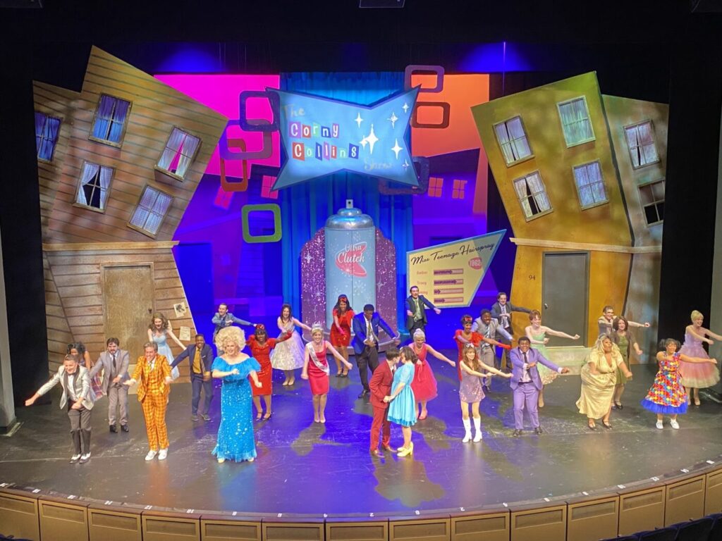 A large ensemble performs on a brightly lit stage with tilted building facades and colorful retro set pieces.