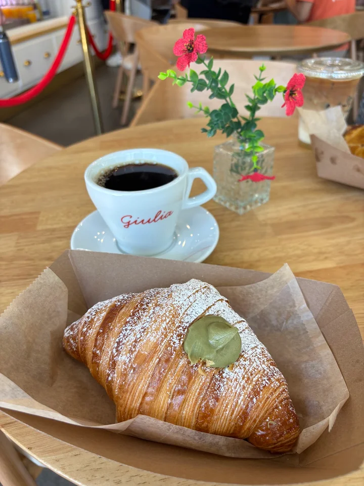 A golden croissant dusted with powdered sugar and green cream filling beside a white Giulia-branded cup of black coffee on a wooden café table.