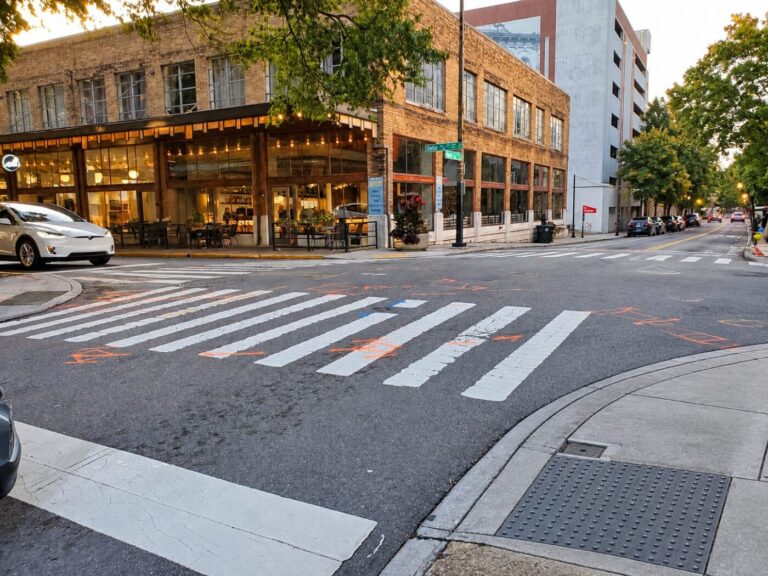 A downtown street corner with a painted crosswalk, brick storefronts and parked cars at dusk.