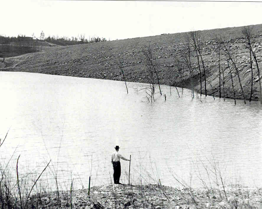 A man stands beside the filling Lake Lanier reservoir, with bare trees in the water and a stone-covered embankment beyond.
