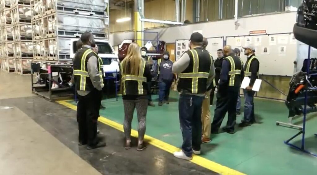 A group of people wearing safety vests stands inside an industrial facility near a vehicle and machinery.