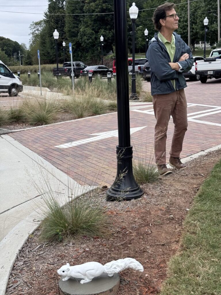 A white squirrel sculpture near a sidewalk lamp post with a man standing nearby.